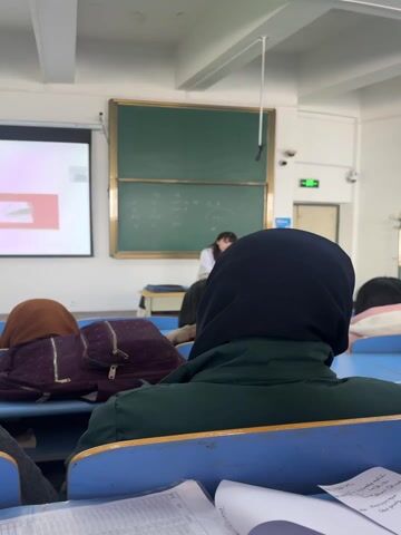 The video shows a classroom scene from the perspective of a student seated towards the back. In the foreground, the back of a student's head is visible, wearing a dark green jacket and a black hijab. On the desk directly in front of the camera, a tablet is open, displaying text related to a multiple-choice question: "The trigger of classical pathway is" with options including "Ag-Ab complex" and "C3b-Surface of pathogen," indicating a subject like immunology or biology.

Further ahead, another student wearing a brown head covering is visible, with a maroon backpack resting on their blue desk. In the front of the classroom, a female teacher, dressed in a white top and blue bottom, is standing near a green blackboard that has some handwritten notes or equations in white chalk. To the left of the blackboard, a projector screen displays a slide with a circular logo and the words "Thank you!" in a red rectangle, suggesting the conclusion of a presentation segment.

The classroom features blue desks and chairs arranged in rows. The ceiling is white with multiple fluorescent lights and what appear to be security cameras mounted near the ceiling. Throughout the video, a female voice, presumably the teacher, can be heard speaking, occasionally using English words like "pathogen." The camera makes slight panning movements, briefly revealing the side profile of another student with long dark hair looking down at their desk. The overall atmosphere is that of an active academic lesson.