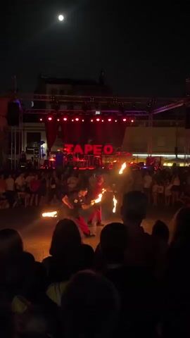 The video shows an outdoor nighttime performance with a stage in the background. Two performers are on a dirt or paved area in front of the stage, surrounded by a crowd of spectators. The performers are holding torches that are on fire and appear to be performing a fire-dancing or fire-eating act. The stage is lit with red lights, and a sign with the word "ZAPEO" in red letters is visible above the stage. A full moon is visible in the dark sky. The crowd is watching the performance, and some people are holding up their phones to record. The atmosphere appears to be festive and energetic.