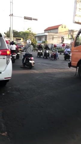 The video shows a street scene in Klurak, Indonesia, likely during the late afternoon or early evening. The lighting suggests a low sun angle. The road is paved and appears to be a main thoroughfare with multiple lanes.

The primary subjects are numerous people riding motorcycles. Most are wearing helmets, and some are carrying passengers or small cargo. The motorcycles are of various models and colors. In the foreground, the rear of a white car is visible, suggesting the camera is mounted on or in a moving vehicle. To the right, the side of an orange truck or delivery vehicle is partially visible. In the background, there are buildings, including what appear to be houses or low-rise commercial structures with red-tiled roofs. Trees and foliage are also present.

The activity is traffic, with many motorcycles navigating the road. Some are in motion, while others may be stopped at an intersection, though this is not definitively clear. The overall impression is one of busy, everyday traffic.

Notable details include the abundance of motorcycles, a common mode of transport in Indonesia. The weather appears to be clear, with no visible rain or overcast skies. The time of day is suggested by the warm, low light. The location cues are the style of buildings and the prevalence of motorcycles. There are no strong emotional cues evident in the captured footage; it's a straightforward depiction of traffic.

There is no clearly legible text visible in the video.