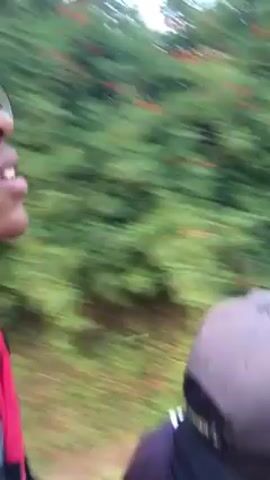 The video shows a person riding a motorcycle on a dirt road. The person is wearing a purple jacket with white stripes and a black baseball cap. There is another person riding behind them. The vegetation is green on either side of the road. The motorcycle has a digital speedometer and rearview mirrors. The road is sandy.