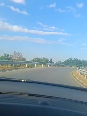 The video is a first-person view from inside a car driving on a road with guardrails on both sides. The road curves to the right. The sky is blue with scattered white clouds. Trees and bushes line the side of the road.