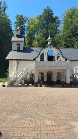 The video shows a white, domed church or monastery. The building has multiple entrances, staircases, and arched windows. There are gold domes on top of some of the structures. The building is set against a backdrop of lush green trees and a clear blue sky. The ground in front of the building is paved with brick-like stones. There are potted plants placed around the entrance and along the staircases. One person is visible standing on a balcony or upper level of the building. The camera pans slowly across the facade of the building.