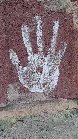 A close-up shot of a white handprint is painted on a reddish-brown brick wall. The wall appears weathered, with some mortar visible. The handprint is stylized, with four fingers clearly defined and the thumb partially visible. The paint is slightly faded and uneven, suggesting it has been there for some time. The camera slowly pans to the right, revealing a dark crevice between two bricks on the left side of the frame.