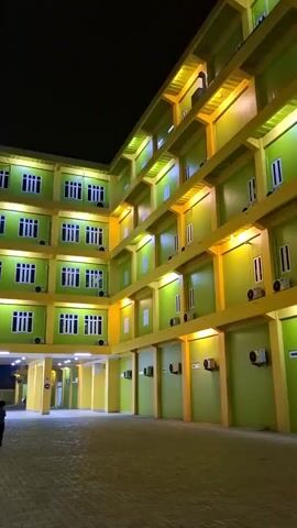 This media file captures the exterior of a modern, multi-story building at night, likely a hotel or dormitory, in Lampeuneurutblang, Indonesia. The building features a striking lime green facade contrasted with bright yellow trim and structural elements. Numerous windows, each with white frames and visible air conditioning units below, are evenly spaced across each level.

The building is illuminated by warm, yellow artificial lights that highlight the architectural features and create a welcoming glow against the dark night sky. The perspective of the camera suggests the viewer is in an open courtyard or parking area in front of the building, paved with interlocking bricks or similar material.

The primary activity suggested is the nightly operation of the establishment, with its lights on and air conditioners running. No people are visible, and the scene is quiet and still, conveying a sense of calm and order.

Notable details include the time of day being night, indicated by the dark sky and artificial lighting. The weather appears clear. Location cues are subtle, but the text on the ambulance in the lower right corner provides a significant clue. The text on the ambulance reads "AMBULANCE" and below that, in smaller print, "AKSU PUTRI BIDADARI" and "Jl. SOKARNO HATTA KM 10 KAB. ACEH BESAR" and "Kesehatan Anda Merupakan Prioritas Utama". This indicates the ambulance belongs to a medical facility named "AKSU Putri Bidadari" and is located on Soekarno Hatta Road, Kilometer 10, in the Aceh Besar Regency, Indonesia. The presence of an ambulance suggests the building might be associated with a hospital or clinic, or that it is in a general vicinity where emergency services are readily available. The overall emotion conveyed is one of modern infrastructure and accessibility.