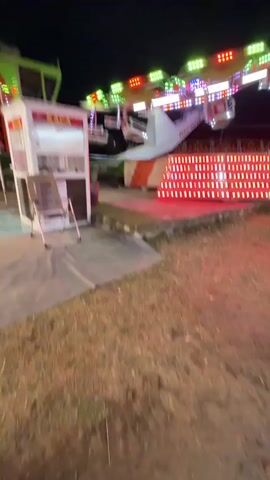 The video shows an amusement park at night. In the foreground, there is dry grass and a grey tarp. To the left, there is a small white booth with the word KACA on it, and a chair in front of it. A portion of an amusement park ride, with lights and what appears to be airplane-shaped cabins, is visible in the center and right of the frame. The ride is illuminated with colorful lights, including red, green, yellow, and white. Further in the background, there are more amusement park rides, lights, and people walking around. The sky is dark. The camera pans slightly to the right, revealing more of the park and a few people walking away from the camera. The scene suggests a lively atmosphere at the amusement park after dark.