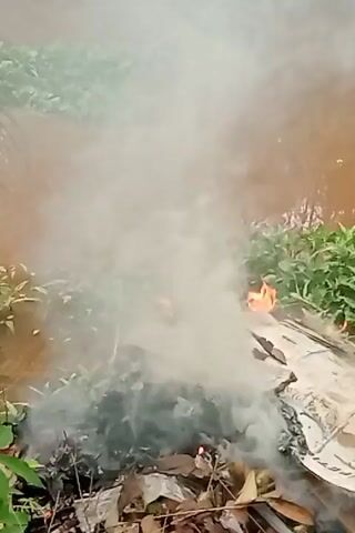 A fire burns in a pile of debris, likely garbage, near a body of water. The fire is producing a thick cloud of white smoke that rises into the air. The debris includes cardboard and dried leaves. Green vegetation is visible on the bank of the water, which appears to be a muddy brown color. The camera is held at a medium distance from the fire.