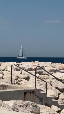 The video shows a sailboat on a calm blue ocean. In the foreground, there are large rocks and a concrete structure with metal railings. The sky is clear and blue.