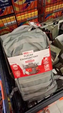 The video shows a hand reaching into a gray backpack. The backpack is branded with a red and white tag that says "MobiPack PLECAK EWAKUACYJNY 21 ELEMENTOW". Several boxes are visible behind the backpack, including boxes of laundry detergent. The scene appears to be in a store aisle.