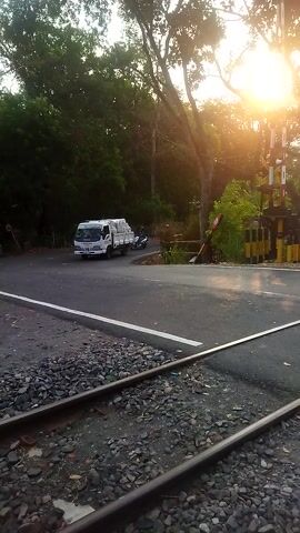 The video shows a truck and a motorcycle at a railroad crossing. The sun is setting, casting a golden light on the scene. Trees line the road, and the railroad tracks run alongside the road. The truck appears to be stopped at the crossing, waiting for a train to pass. The motorcycle is also stopped. The scene is peaceful and quiet.