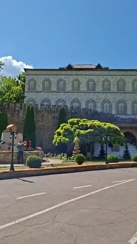 A video captures a sunny day with a clear blue sky and scattered white clouds. In the foreground, a paved road with white lane markings is visible. Beyond the road, a decorative stone wall with battlements forms the boundary of a landscaped area. Several conical evergreen trees and meticulously trimmed spherical bushes are planted along the wall and within the grounds. A fountain with a carved stone centerpiece, possibly depicting a lion or bear, is a focal point in the garden. A lamppost stands near the fountain. On the right side of the video, a building with a grey, textured facade and arched windows can be seen behind the wall. To the left, lush green trees form a dense canopy.

People are seen interacting with the environment. In the first frame, two individuals, one appearing to be a man with a hat and the other possibly a child, are near the fountain. In subsequent frames, two women are seen walking across the road, and then a man and a woman are walking together. Another woman is seen walking away from the camera on the road. The overall impression is a pleasant outdoor setting, possibly a park or a historical site, with visitors enjoying the day.