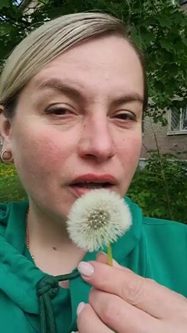 A woman in a green hoodie blows on a white dandelion flower. She is holding the flower with her left hand. She has fair skin and red lipstick. A gold chain is visible around her neck. In the background, there is green foliage and a brick building with a white-framed window.