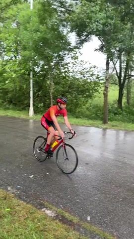 The video shows a cyclist riding a red bicycle on a wet, gray asphalt road. The cyclist is wearing a red and black uniform with "HUB" printed on the shorts. Trees line the road, creating a green backdrop. In the background, more trees and bushes can be seen. The road is wet, indicating recent or ongoing rain. A person with an umbrella walks into the shot, moving toward the camera. There are also yellow cones visible on the road surface.