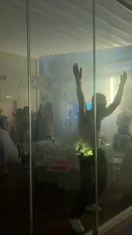 A man with his arms raised is dancing energetically in front of a glass wall during a party. Through the glass, a group of people are visible dancing and socializing in a room filled with smoke and red and white lights. Tables with chairs are set up in the background. The man's dance moves appear enthusiastic and uninhibited, suggesting he is enjoying the music and atmosphere.