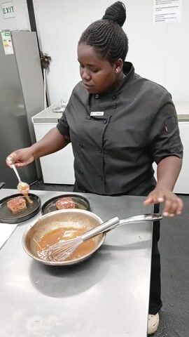 A woman in a black chef's uniform is working at a stainless steel table in a commercial kitchen. She has her hair braided and styled in a bun. Her name tag reads "Anita". She is carefully spooning sauce from a metal bowl onto two pieces of what appear to be browned cakes or meat on black plates. A whisk rests in the bowl of sauce. Another person hands her a small metal container of greens. The background includes a refrigerator, an exit sign, and a sign with multiple lines of text. The floor appears to be tiled.