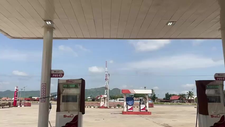 This media file captures a wide shot of a gas station in Pasere, Nigeria. The scene is set under a covered canopy supported by white pillars, illuminated by fluorescent lights. Several gas pumps are visible, some with brown coverings and digital displays showing "Shafa" and "PMS 3" or "PMS 2". One central pump island has a red and white structure and appears to be unused.

In the background, a communication tower stands tall against a bright, partly cloudy sky. Beyond the gas station, a collection of buildings, some with red roofs, are scattered across the landscape, suggesting a populated area. Hills or mountains are visible in the distance.

Two people are present in the frame. One individual is seated on a fuel pump, and another stands next to a dark-colored car, seemingly waiting or conversing. The overall atmosphere appears calm and casual. The time of day is daytime, with clear, sunny weather. The setting is an outdoor public space, likely a functional commercial establishment. Visible text includes brand names like "Shafa" and fuel type indicators "PMS".