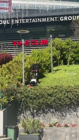 A man is seen mowing a grassy area in front of a building labeled "ISTINYE ENTERTAINMENT GROUP" and "THE CLUB". The man is wearing sunglasses, a black jacket, and red gloves. He is pushing a white and black lawnmower. The area includes bushes, trees, and a trash can. The building has a modern design with horizontal lines. The weather appears sunny and clear.