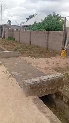A drone shot pans across a dirt road with a concrete drainage ditch running alongside it. On the other side of the ditch is a cinder block wall, topped with what appears to be a metal fence. Beyond the wall, there are some trees and houses. A yellow jerrycan and some metal sheeting are leaning against the fence. The area appears to be uncultivated, with some sparse vegetation growing in the dirt. The sky is overcast.