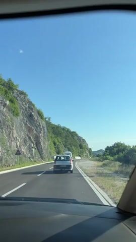The video shows a POV shot from inside a vehicle driving on a road. The road is paved and has white lane markings. On the left side of the road is a steep, rocky hill covered in green vegetation. On the right side of the road, there is a gravel shoulder and more vegetation. Several cars are visible on the road ahead, all traveling in the same direction. The sky is clear and blue. The overall scene suggests a drive through a mountainous or hilly landscape on a sunny day.