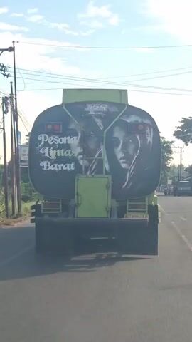 A rear view of a green tanker truck driving on a paved road. The truck has a large mural on its back showing two faces, one on the left and one on the right, with text that reads "PESONA LINTAS BARAT" in white, stylized lettering. The truck has a yellow license plate. The road appears to be in a rural or suburban area, with trees and some buildings visible on the sides. The sky is blue with some clouds. There are other vehicles in the distance on the road.