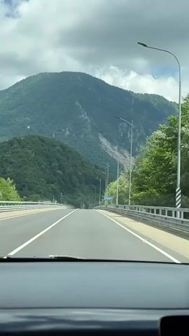 The media file shows a point-of-view shot from inside a car driving on a highway. The highway has multiple lanes and is bordered by metal guardrails. On the right side of the road, there are streetlights and trees. In the distance, a large, green, forested mountain dominates the landscape. The sky is partly cloudy, suggesting it is daytime. The weather appears to be mild. The overall scene conveys a sense of travel and journey through a mountainous region.

A blue sign is visible on the right side of the road with white text. While the text is somewhat blurry due to the car's motion and distance, it appears to be in Cyrillic script. Based on the provided location context of Adlerskiy, Russia, it is likely a directional or informational sign related to a town or landmark in that area. The sign seems to read "р. МЗЫМТА" which is likely referencing the Mzymta River, a significant river in the Adlerskiy district. There are no people clearly visible in the shot, emphasizing the focus on the scenery and the act of driving. The atmosphere is serene and picturesque, with the vastness of nature contrasting with the man-made highway.