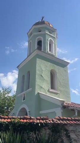 The video begins with a close-up of a light green bell tower. The tower has arched windows and is topped with a copper dome. The camera then pans to the right, showing a stone wall with red tile roofing, followed by a large green tree with leaves and then several tall pine trees. The video continues to pan right, revealing a narrow road bordered by trees and vegetation. The sky is bright blue with some scattered white clouds. There are no people or animals visible in the video.