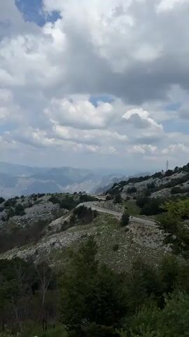This is a video capturing a panoramic view of a mountainous landscape in Žanjev Do, Montenegro. The scene is dominated by rolling hills and rugged mountains, some of which are covered in a sparse, rocky terrain with patches of green vegetation and trees. A winding asphalt road is visible, snaking its way up one of the slopes, hinting at a journey or exploration. In the distance, a vast expanse of mountains fades into a hazy blue, suggesting a great depth to the landscape. The sky is filled with dramatic, white and gray clouds, indicating a dynamic weather system, perhaps before or after a storm, or simply a cloudy but bright day. The lighting suggests it is daytime, possibly late morning or afternoon, with the sun filtering through the clouds, casting soft shadows and highlighting the textures of the terrain.

The overall mood is one of natural grandeur and a sense of being in a remote, untouched place. The absence of people and urban elements emphasizes the wilderness and the scale of the natural environment. The rough, textured mountainsides, the hardy vegetation clinging to the slopes, and the vast, layered horizon all contribute to a feeling of awe and the wild beauty of the Montenegrin terrain. The video seems to be a static shot, allowing the viewer to absorb the expansive scenery. There is no visible text within the video frame.