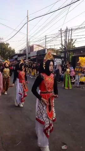 A group of young women in traditional attire are marching down a street. They are wearing elaborate headdresses and colorful costumes, including patterned skirts and decorative vests. Some of the women are wearing headscarves. They appear to be participating in a parade or cultural event. The street is lined with buildings and power lines, and there are spectators watching the procession. The overall atmosphere seems festive.