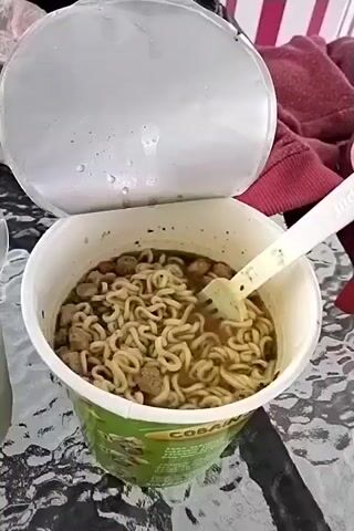 A close-up video shows a cup of instant ramen noodles being stirred and lifted with a white fork. The cup is white with a green label that reads "COBAIN". The lid is open and partially peeled back. The noodles are golden brown and appear to be in a broth. There are other ingredients in the cup, possibly pieces of meat or vegetables. In the background, a person's hand is visible, holding the fork. A partially visible drink in a clear plastic cup is to the left. The surface the cup is on appears to be a glass table with a textured pattern. The background is blurry but suggests an indoor or outdoor setting with some fabric items, possibly clothing.