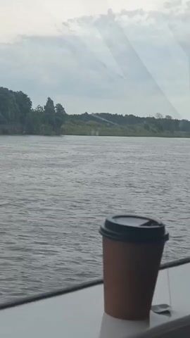 A disposable coffee cup with a black lid sits on a white ledge next to a window. Beyond the window, a wide, calm river flows under an overcast sky. The opposite bank of the river is lined with lush green trees and vegetation. A small, silver spoon is visible on the ledge next to the cup. The scene appears to be from a moving vehicle, possibly a boat or a train, as indicated by the slight motion blur in the background.