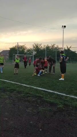 The video depicts a group of children and adults on a grassy sports field, likely a soccer field, given the presence of a goal in the background and the attire of some of the individuals. The children, dressed in various colored athletic wear, appear to be engaged in some form of activity or training. Several adults are also present, some of whom seem to be coaching or supervising the children. The sky is overcast, suggesting it is either early morning or late afternoon. The overall atmosphere appears to be one of organized sports practice.