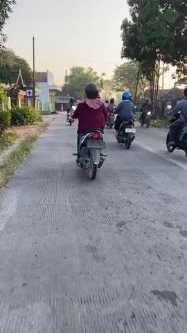 The video shows a group of people riding motorcycles on a paved road. The motorcycles are moving in the same direction, following one another. The riders are wearing helmets, and some are carrying backpacks. The environment appears to be a rural or suburban area with trees and buildings visible on the sides of the road. The lighting suggests it is either early morning or late afternoon. One motorcycle in the foreground is red, and another in the background is black. The road has concrete barriers on the edges.