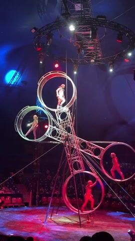 This video shows a circus performance with four acrobats in metallic, futuristic-looking costumes. They are performing inside a large, intricate metal contraption that resembles a rotating, multi-ringed structure. Each ring appears to be independently controllable, allowing the acrobats to move within and between them. The performance takes place in a dimly lit circus tent, with spotlights illuminating the performers and the structure. The audience is visible in the background, seated in rows of red chairs. The acrobats are shown running, balancing, and interacting within the spinning rings, demonstrating remarkable agility and coordination. The contraption itself is suspended from the ceiling of the tent and is a complex arrangement of interconnected rings and a central support structure. The lighting shifts between blue and red hues, enhancing the dramatic effect of the performance.