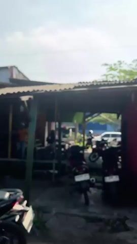 The video shows an outdoor scene in daylight with a slightly overcast sky. The foreground is dominated by the blurry image of the back of a motorcycle. In the midground, there is a covered outdoor area that appears to be a shop or a workshop. Several motorcycles are parked under the shelter. To the left, a person in an orange shirt and blue jeans is visible near what looks like a counter or a table. Further back, other people and more motorcycles can be seen. The structure has a corrugated metal roof supported by posts. To the right of the covered area, a red wall is visible. In the background, there is more greenery and a white vehicle. The overall impression is a busy, casual outdoor commercial space.