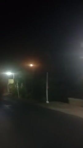 The video is a first-person perspective recording taken at night on a street. There are streetlights illuminating the scene, and some buildings are visible along the side of the road. The road appears to be paved and wet, suggesting recent rain or mist. In the foreground, a motorcycle is visible, and the camera appears to be mounted on it, as it is moving. Two people are riding on the motorcycle, with the person in the back appearing to be looking around. Further down the road, a yellow truck is parked or moving slowly. The overall atmosphere is dark and slightly hazy due to the low light conditions and potentially fog or mist.
