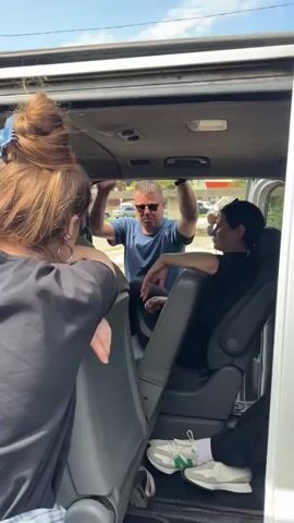 The video shows the interior of a van. In the foreground, the back of a woman with her hair in a bun and wearing a black shirt and a plaid skirt is visible. She is looking into the van. Inside the van, a man in a blue t-shirt and sunglasses is standing and gesturing with his hands, facing two women. One woman is seated on the right side of the van, wearing a black t-shirt and black pants, and looking at the man. Another woman is seated behind her, partially visible. The man appears to be speaking to the seated women. Outside the van, through the open doors, a sunny day and some buildings are visible. There are also some balloons in the background. The focus of the video is on the interaction between the people inside the van.