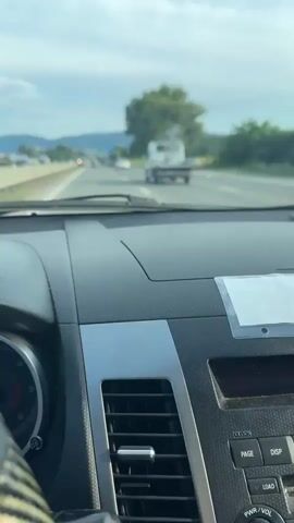 This is a first-person perspective video taken from inside a car driving on a highway. The dashboard of the car is visible in the foreground, with a speedometer and air vents. The road ahead is a multi-lane highway with other vehicles driving in the same direction. There are trees and hills in the background. The sky is cloudy with patches of blue. A white flatbed truck is visible in front of the car. A speed limit sign is visible on the left side of the road. The video captures the experience of driving on an open road, with the scenery changing as the car moves forward.