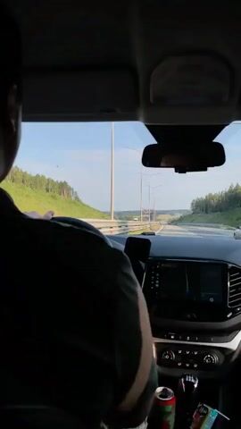 The video shows the interior of a car from the perspective of a passenger. A person is driving, viewed from behind. The dashboard displays a GPS navigation system. Outside the car, a highway with trees and a blue sky is visible. A can of soda and other items are on the center console. The car appears to be in motion on a highway.