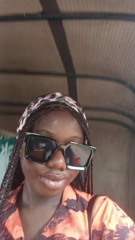A woman with dark skin and braided hair is recording a video of herself inside a moving vehicle. She is wearing sunglasses and a colorful orange and black shirt. The vehicle appears to be a rickshaw or tuk-tuk, with a metal frame visible above her. She touches her forehead with her hand. There is a blurry green and white sign behind her. The video seems to be taken during the daytime in an outdoor setting.