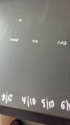 A dark, flat surface is marked with white chalk. Writing in chalk includes "x", "шар", "пл", "нез", "шар", and "пл". Below this, numbers are written, starting with "3/10", then "4/10", "5/10", "6/10", and "7/10". A metallic object, appearing to be a ball bearing or similar small sphere, is being held by a human finger and thumb, positioned near the chalk marks. Another chalk mark, resembling a lowercase "r", is visible above a small pile of white granular material. The camera angle is downward, looking at the surface.