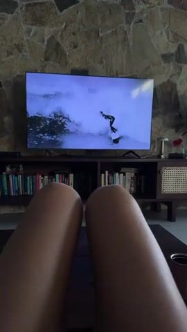 The video shows a first-person perspective of someone relaxing indoors, with their legs in the foreground and a television screen in the background. The television displays a surfing video of a surfer riding a wave. The room has a stone wall behind the television, and a wooden shelf unit filled with books and decorative items. The lighting is dim, with the primary light source coming from the television screen. The overall atmosphere is one of leisure and relaxation.