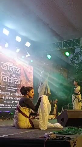 A group of women in traditional Indian clothing are on a stage performing. The woman in the foreground is kneeling and holding a white cloth. She is wearing a white sari with a dark blouse. The other women are also kneeling behind her, some with their heads bowed. The stage is lit with spotlights and smoke. There is a large screen behind the performers with text in an Indian language. The overall setting appears to be an outdoor cultural event.
