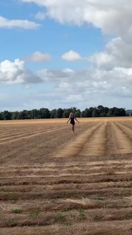 A young blonde woman in a dark swimsuit runs through a dry, harvested field of flax. The field is laid out in long, straight rows of dried flax. She is running away from the camera towards a line of trees in the distance. The sky is blue with scattered white clouds. The sun is shining. The video is shaky and appears to be filmed with a handheld device.