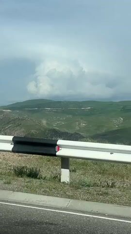 The video captures a wide, scenic view of a mountainous landscape, likely taken from a moving vehicle. In the foreground, a white and black guardrail is visible, with a small red and white marker on its white section. Beyond the guardrail, the scene opens up to expansive, rolling green hills and mountains under a partly cloudy sky. The terrain appears rugged in places, with some patches of lighter, possibly rocky or dry ground interspersed with lush green vegetation. Large, white, puffy clouds dominate the sky, suggesting a bright but potentially changeable weather day. The overall impression is one of vast natural beauty. The setting is consistent with a roadside view in a rural or mountainous region, specifically Yeraskh, Armenia, as provided in the context. There is no discernible text on the guardrail marker.