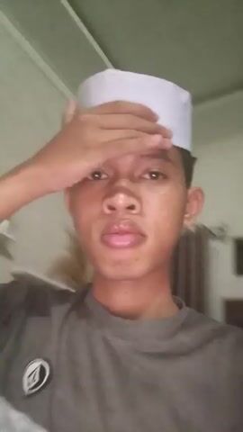 The video shows a young person wearing a white kufi and a dark t-shirt. The person is indoors, with curtains and a bed visible in the background. The person is touching their head and face, and appears to be looking directly at the camera. The lighting is dim.