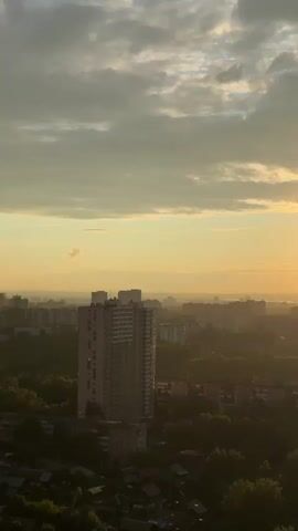 The video shows a cityscape at sunset. The sun is low on the horizon, creating a golden glow over the city. There are several tall buildings and trees visible in the foreground. The sky is filled with clouds, some of which are illuminated by the setting sun. A lens flare is visible near the sun.