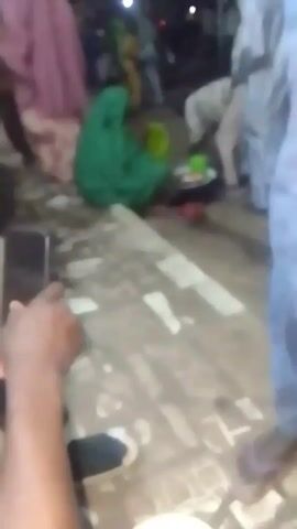 The video shows an outdoor scene at night. Several individuals are visible, some wearing traditional garments. One person in a green garment is kneeling. There are dishes on the ground, and some individuals appear to be eating. The ground is uneven and has some debris on it. The video appears to be taken with a handheld device. Due to the quality of the video, many details are blurry and indistinct.