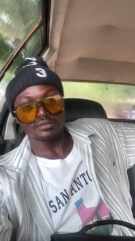 The video shows a man inside of a vehicle. He is wearing a black beanie, yellow sunglasses, a white t-shirt with "SAN ANTONIO" printed on it, and a gray and white striped shirt. The man is looking at the camera with a slight smile. It appears to be raining outside the vehicle. At the beginning of the video, there is a sound similar to a car horn.