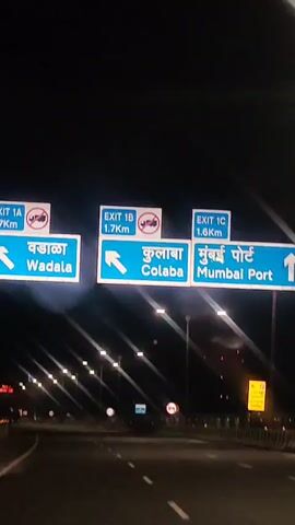 A nighttime shot captures a series of overhead road signs. The signs are blue with white lettering, some featuring directional arrows and place names in both English and what appears to be Marathi. Specifically visible are signs for "Wadala", "Colaba", and "Mumbai", with corresponding distances indicated, such as "1.7 Km" and "1.6 Km". Red and white prohibition signs, indicating no entry for certain vehicles, are also present. Below the signs, streetlights illuminate a multi-lane road, suggesting the footage is from a moving vehicle on a highway or major thoroughfare. The background is dark, consistent with nighttime conditions.