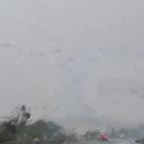 This video captures a view through a vehicle's windshield during a rainstorm. The windshield is wet with raindrops, distorting the view of the outside environment. The scene appears to be a highway with several vehicles visible. In the foreground, a grey barrier runs horizontally across the lower portion of the frame. Beyond the barrier, a road is visible with traffic. On the left side, a white vehicle is seen, and further down the road, a red taillight from another vehicle is illuminated, indicating it is likely moving. Trees and vegetation are present on the sides of the road. The sky is overcast and gray, contributing to the overall dim lighting conditions and low visibility characteristic of a rainy day. The movement of the vehicle causes slight motion blur, further emphasizing the sense of travel.