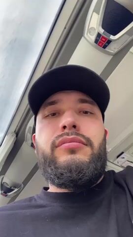 A close-up shot features a man with a beard, wearing a black t-shirt and black baseball cap. He glances slightly to his left and then to his right, then subtly smiles. He appears to be inside a bus or similar transport vehicle, as suggested by the interior lighting and ceiling fittings above him. A window is visible in the background, showing an overcast sky. The camera angle is looking up at him slightly.